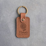 Caramel leather keychain debossed with a sunflower above Nana's and GARDEN, on gray concrete