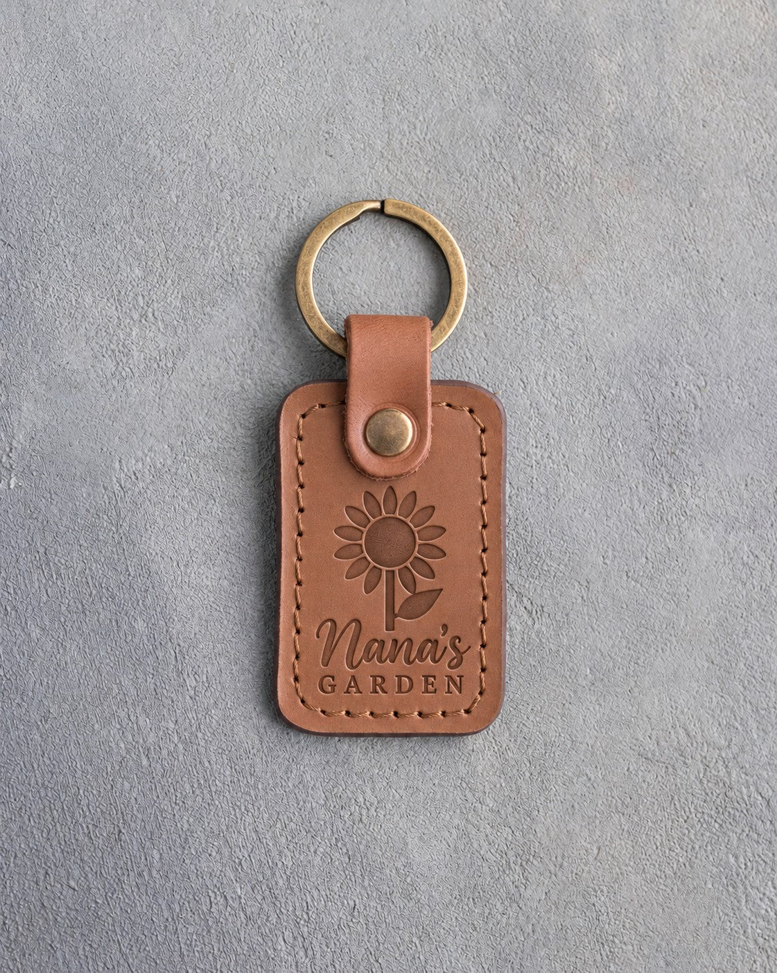 Caramel leather keychain debossed with a sunflower above Nana's and GARDEN, on gray concrete