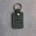 Forest green leather keychain debossed with a mountain range above Parks and 27 OF 63, on gray concrete