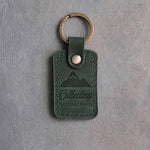 Forest green leather keychain debossed with a mountain range above the words Collecting and NATIONAL PARKS, on gray concrete