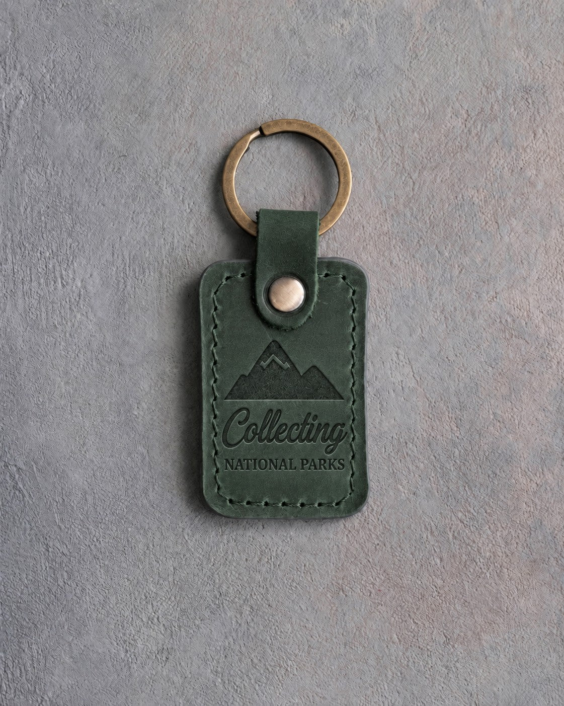 Forest green leather keychain debossed with a mountain range above the words Collecting and NATIONAL PARKS, on gray concrete