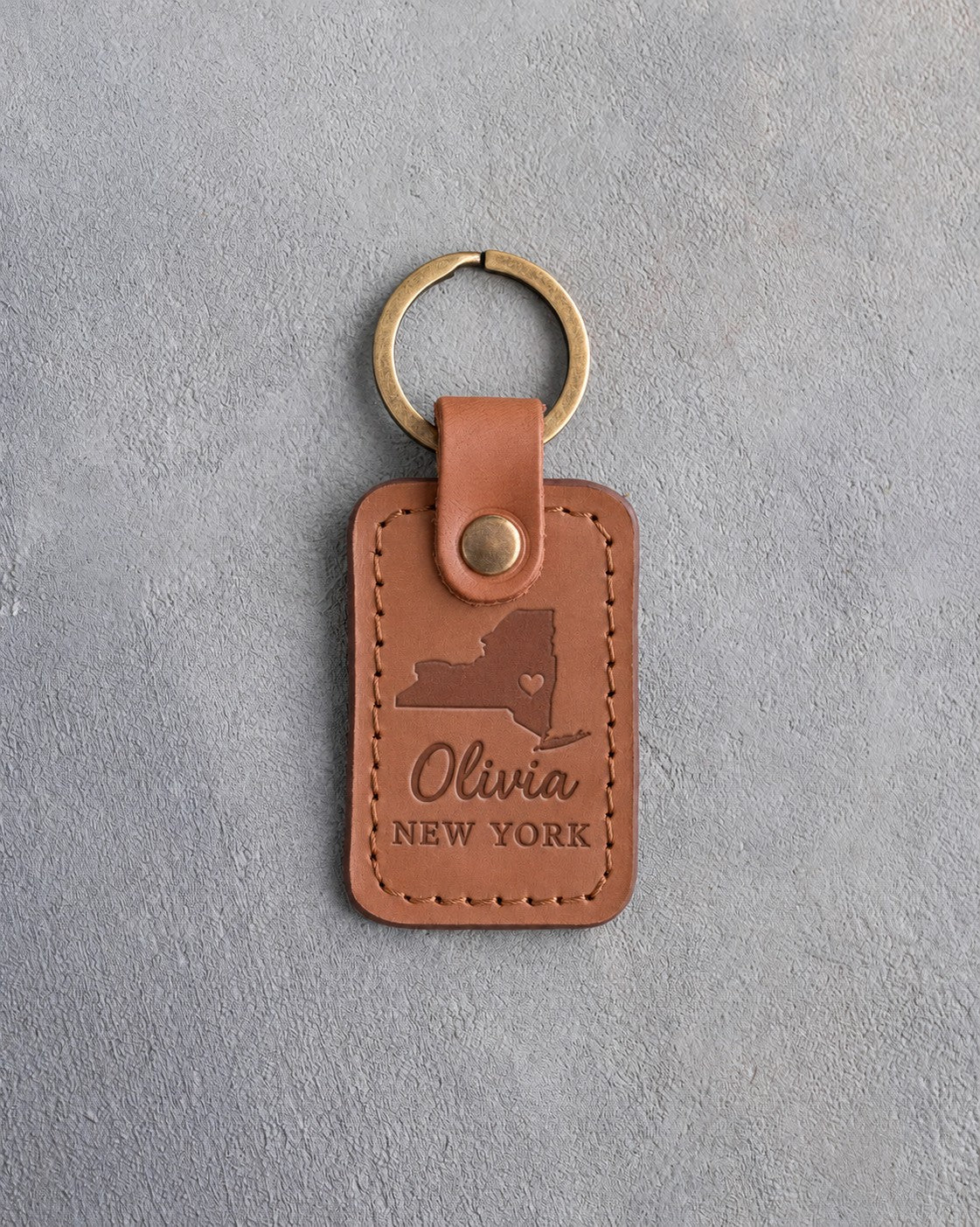 Caramel leather keychain debossed with a New York State silhouette and heart above the name Olivia and NEW YORK, on gray concrete