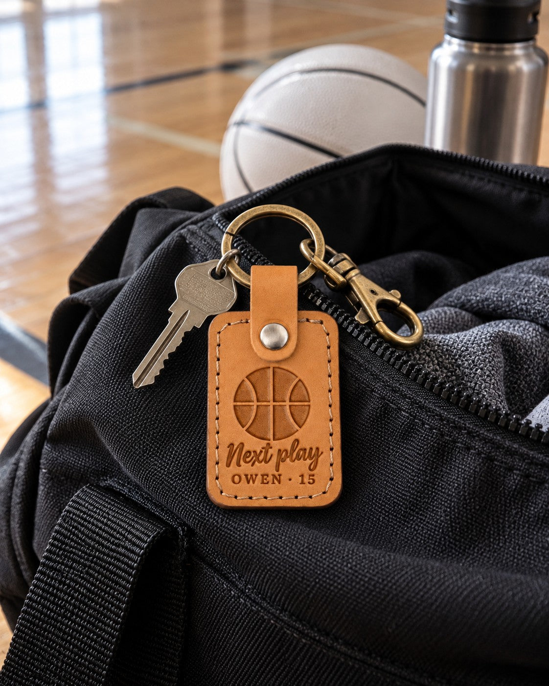 Next Play keychain in golden brown leather clipped to a player's gym bag on a hardwood court