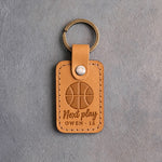 Golden brown leather keychain debossed with a basketball and the words Next play OWEN 15, on gray plaster