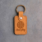 Golden brown leather keychain debossed with a basketball and the words Next play, on gray concrete