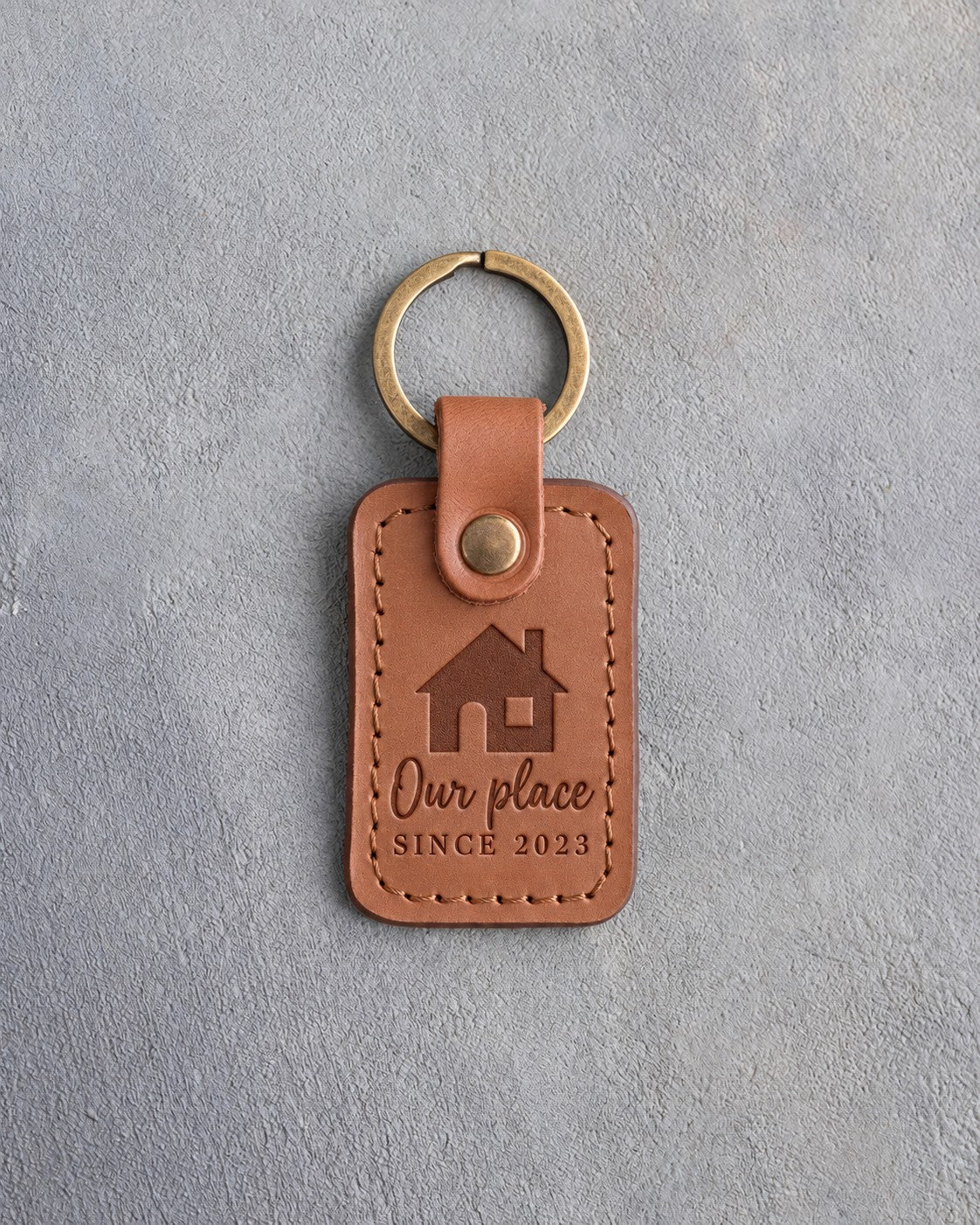Caramel leather keychain debossed with a gable-front house above Our place and SINCE 2023, on gray concrete
