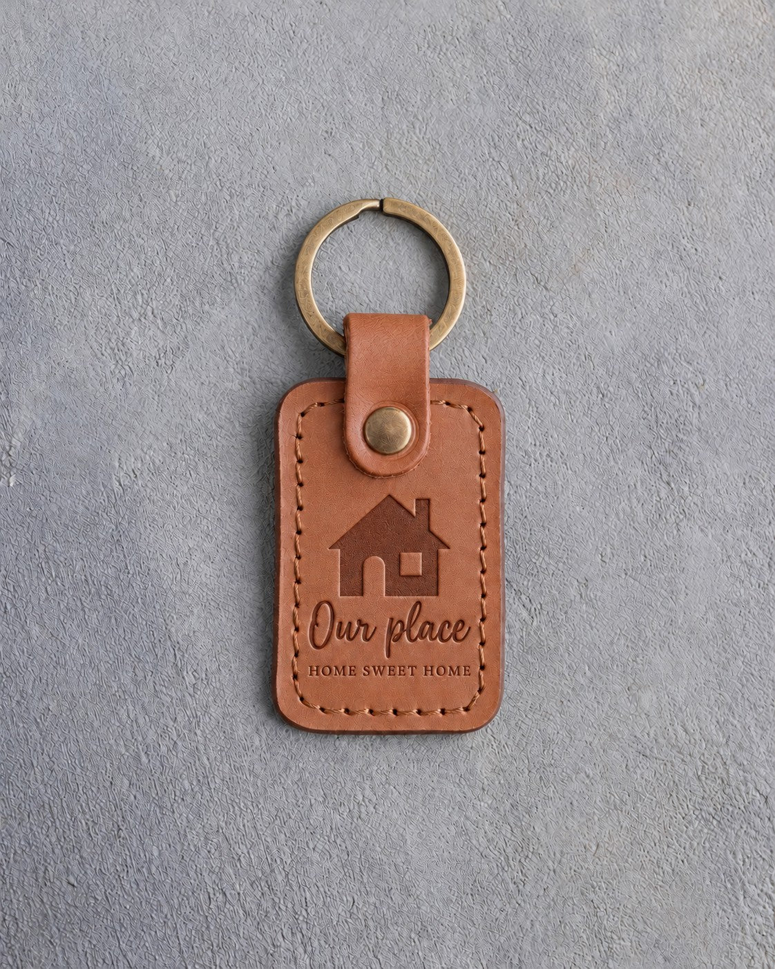 Caramel leather keychain debossed with a gable-front house above the words Our place and HOME SWEET HOME, on gray concrete