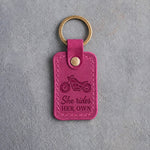 Fuchsia pink leather keychain debossed with a motorcycle above the words She rides and HER OWN, on gray concrete