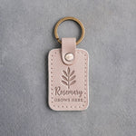 Gray sand leather keychain debossed with a rosemary sprig above Rosemary and GROWS HERE, on gray concrete