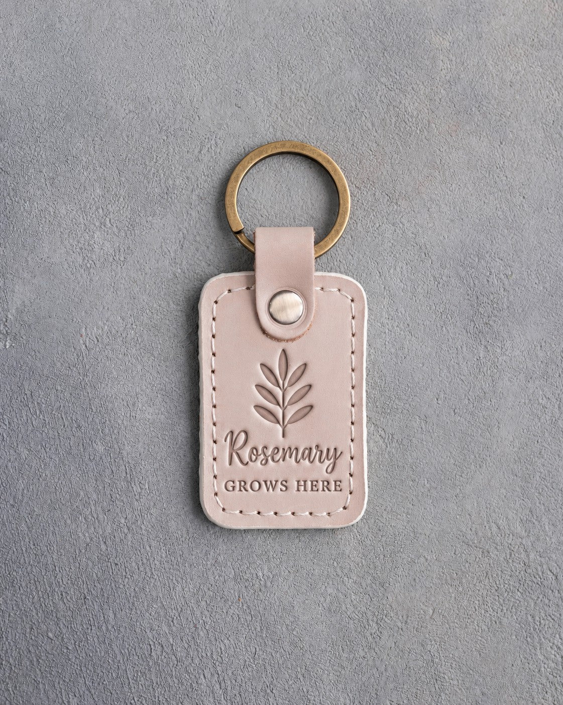 Gray sand leather keychain debossed with a rosemary sprig above Rosemary and GROWS HERE, on gray concrete
