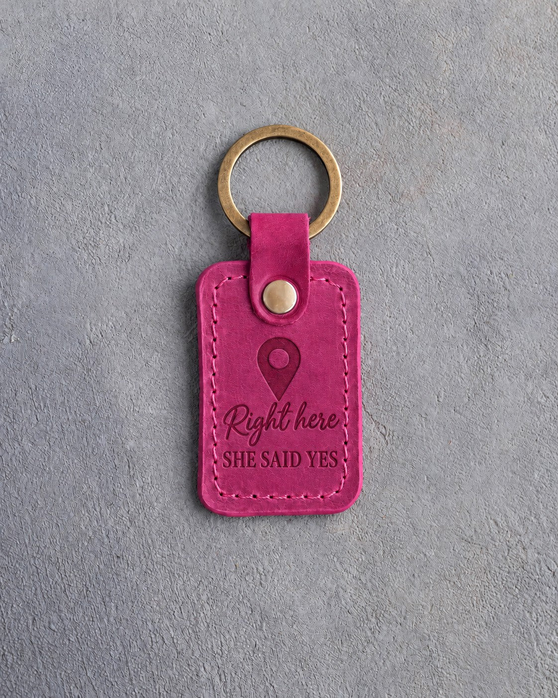Fuchsia pink leather keychain debossed with a map pin above the words Right here and SHE SAID YES, on gray concrete