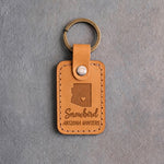 Indian Summer tan leather keychain debossed with an Arizona silhouette and a heart at Scottsdale above the words Snowbird and ARIZONA WINTERS, on gray concrete