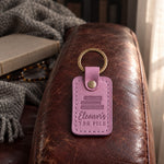 Lilac Eleanor's TBR PILE keychain on the arm of a leather armchair above a closed book and reading glasses