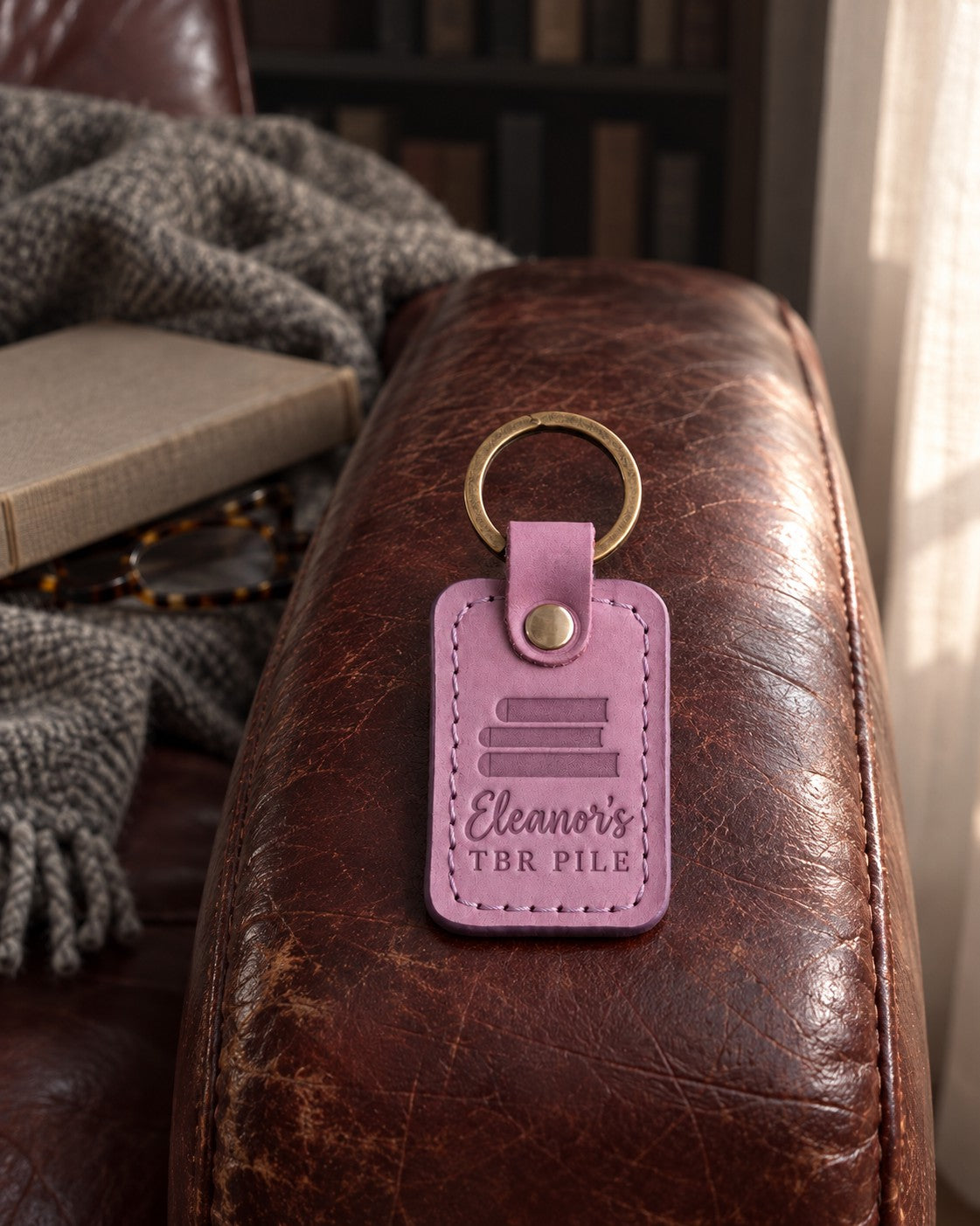Lilac Eleanor's TBR PILE keychain on the arm of a leather armchair above a closed book and reading glasses