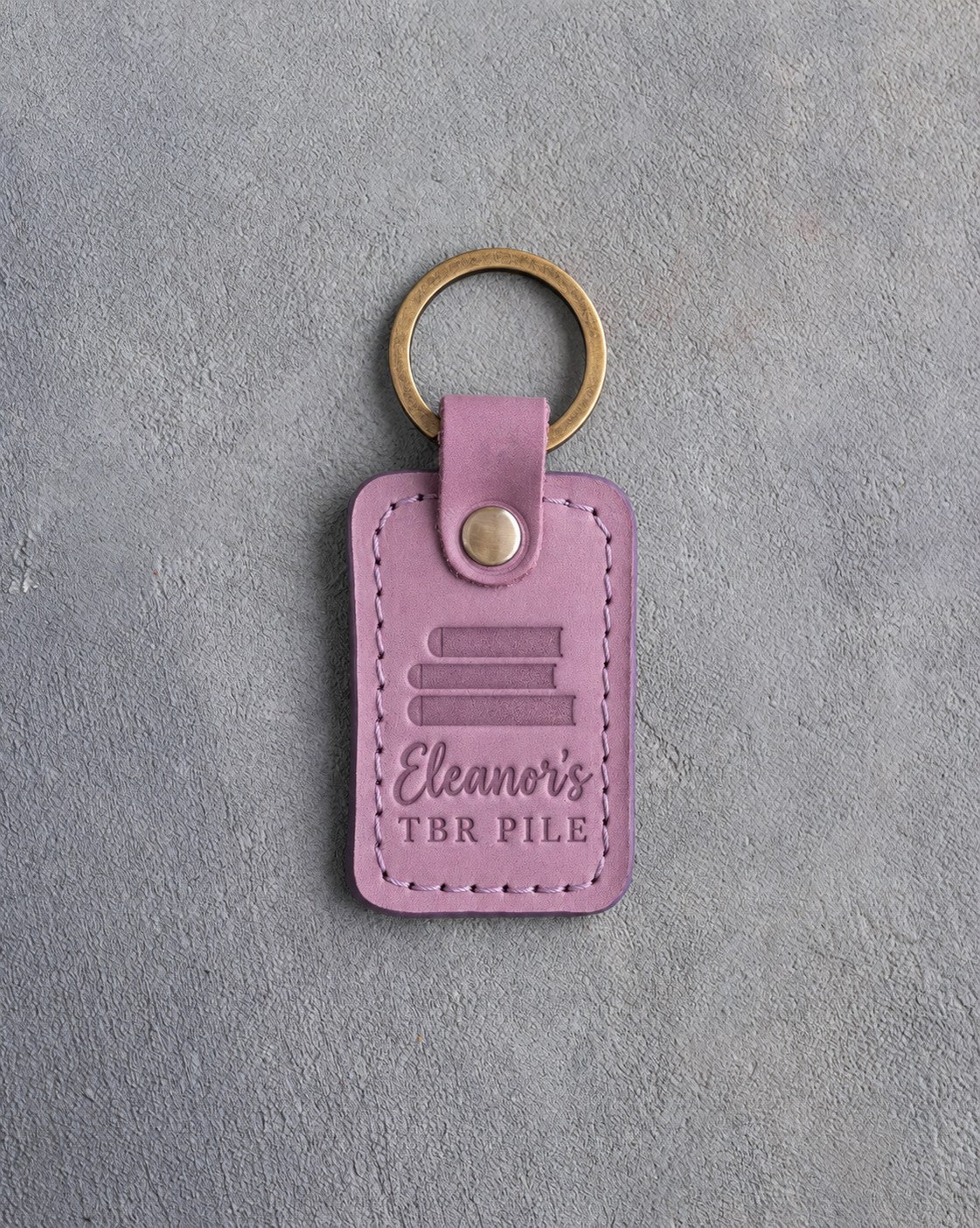 Lilac leather keychain debossed with a pile of three books above Eleanor's and TBR PILE, on gray concrete