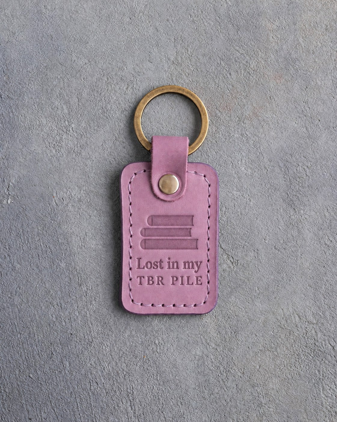 Lilac leather keychain debossed with a pile of three books above Lost in my and TBR PILE, on gray concrete