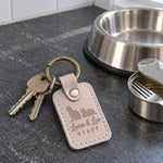Luna & Leo STAFF keychain in pale gray leather beside two cat food bowls on a kitchen floor