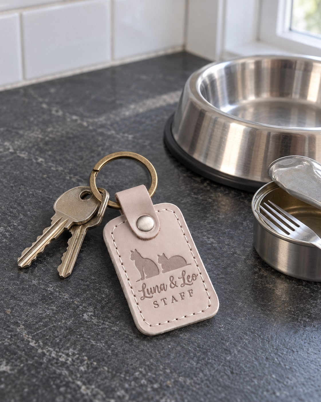 Luna & Leo STAFF keychain in pale gray leather beside two cat food bowls on a kitchen floor
