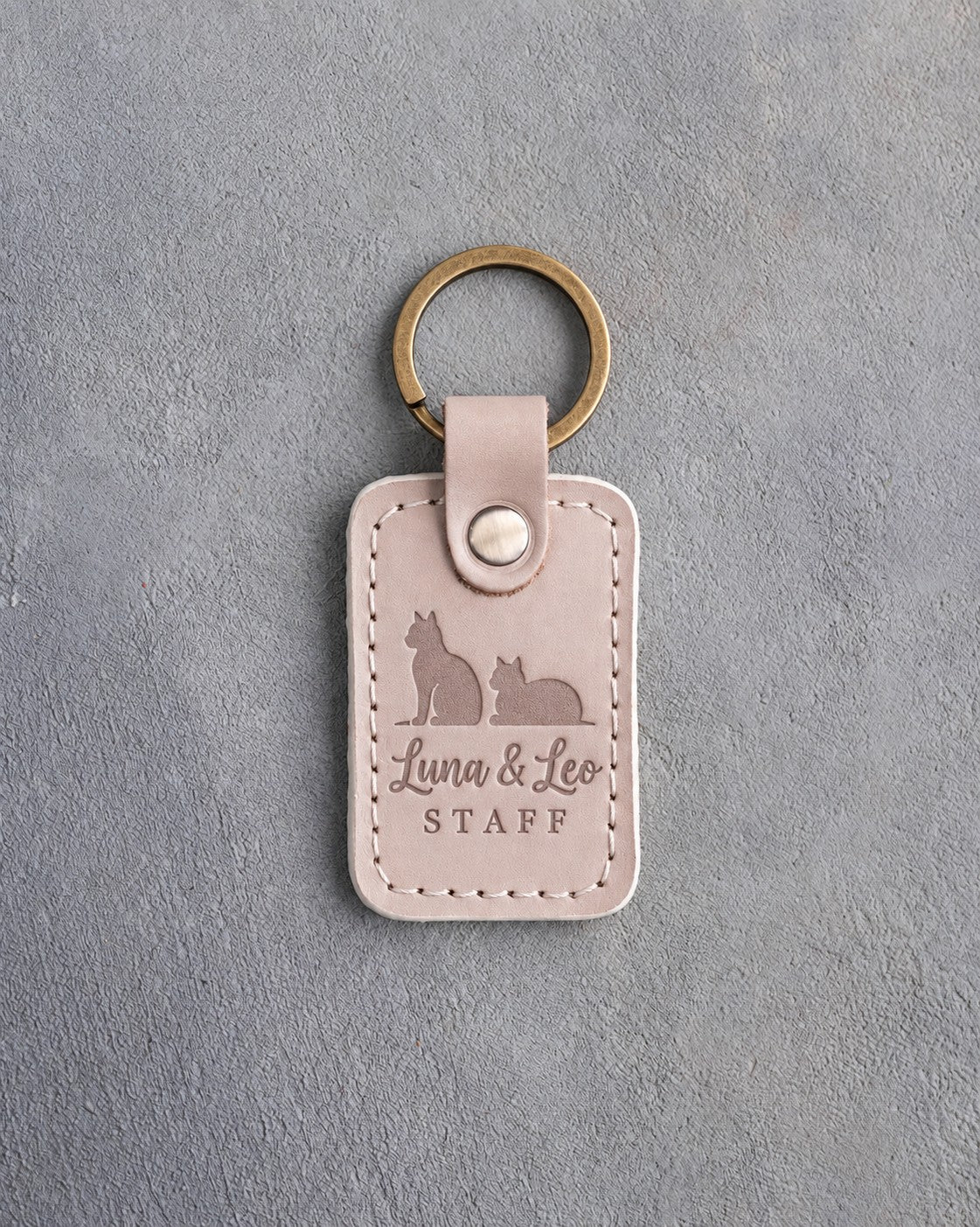 Gray sand leather keychain debossed with a sitting cat beside a loafing cat above the names Luna & Leo and STAFF