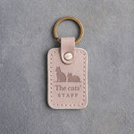 Gray sand leather keychain debossed with a sitting cat beside a loafing cat above The cats' and STAFF, on gray concrete