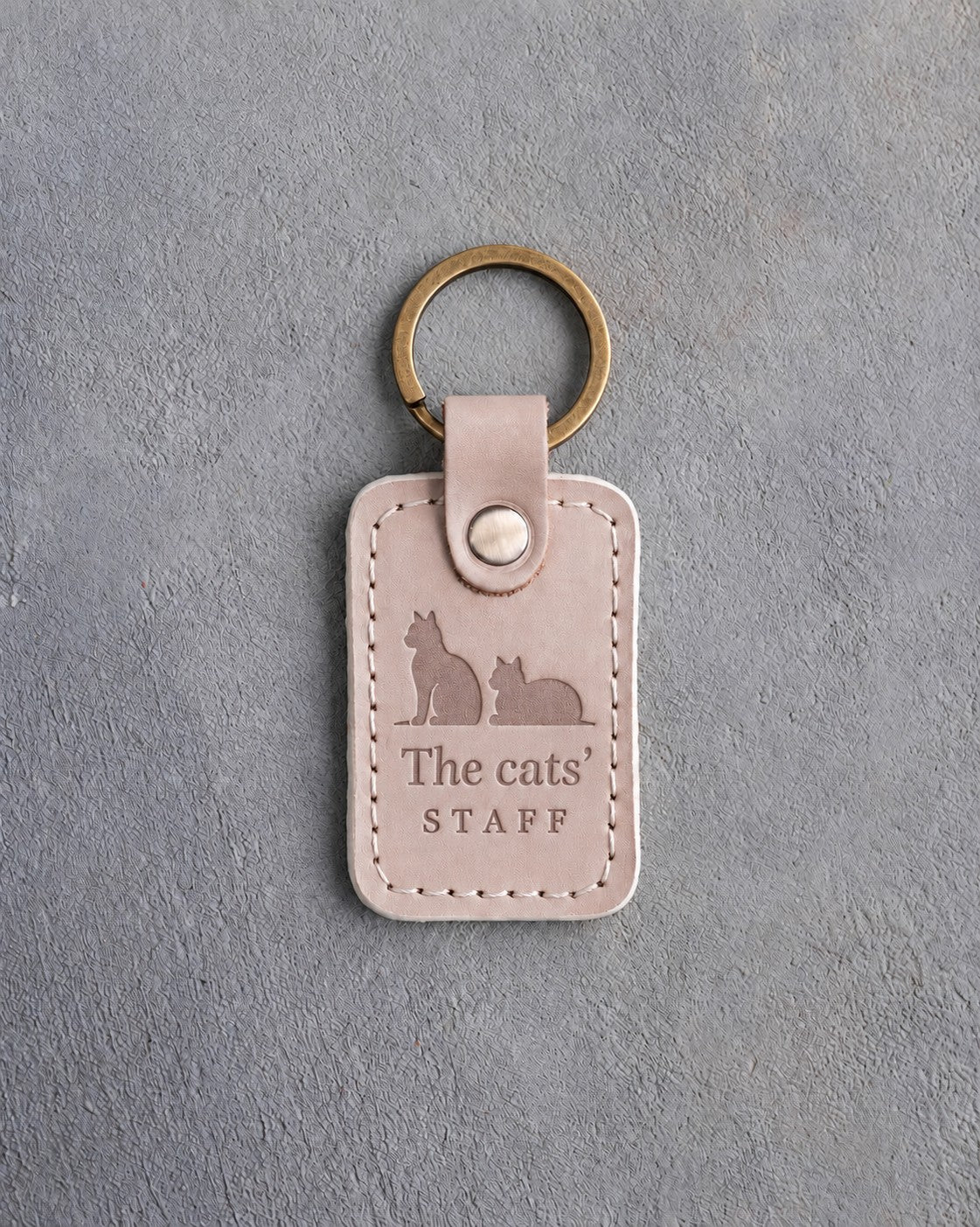 Gray sand leather keychain debossed with a sitting cat beside a loafing cat above The cats' and STAFF, on gray concrete