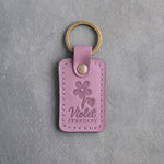 Lilac leather keychain debossed with a violet above Violet and FEBRUARY, on gray concrete
