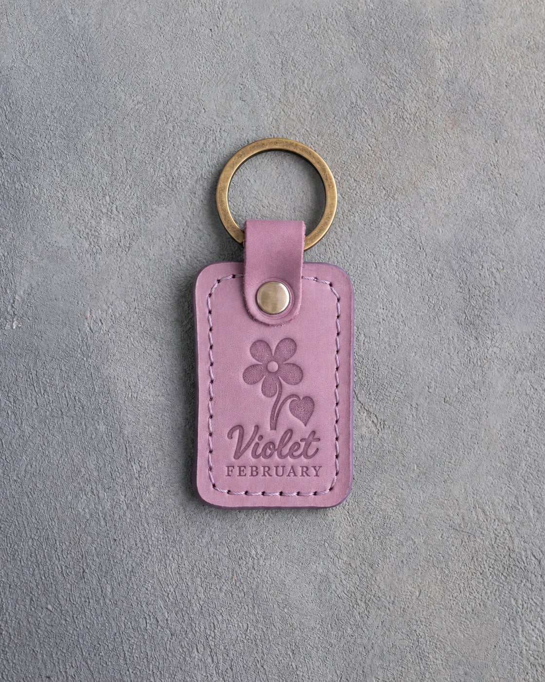 Lilac leather keychain debossed with a violet above Violet and FEBRUARY, on gray concrete