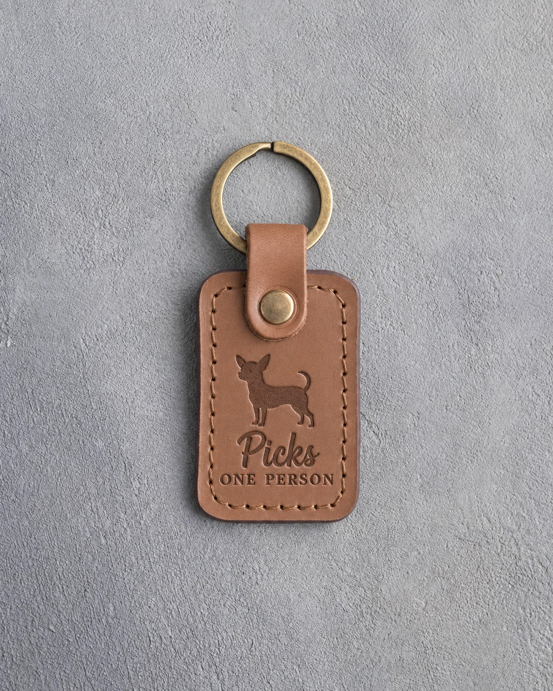 Caramel leather keychain debossed with a chihuahua silhouette and the words Picks ONE PERSON, on gray concrete