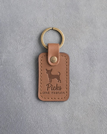 Caramel leather keychain debossed with a chihuahua silhouette and the words Picks ONE PERSON, on gray concrete