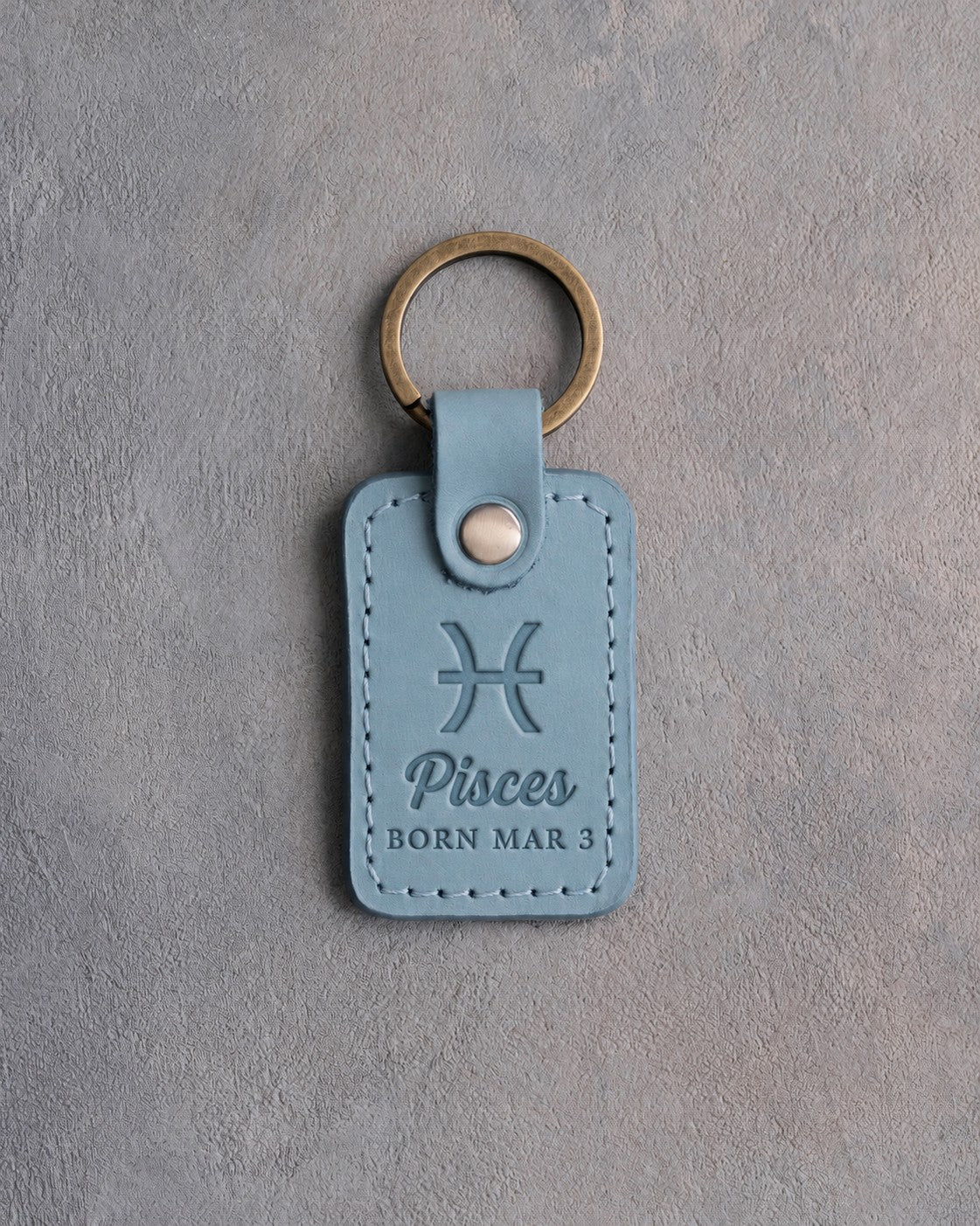 Turquoise leather keychain debossed with the Pisces zodiac glyph above Pisces and BORN MAR 3, on gray concrete