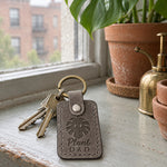 Gray Plant Dad keychain on a wooden shelf beside a large monstera in a ceramic pot