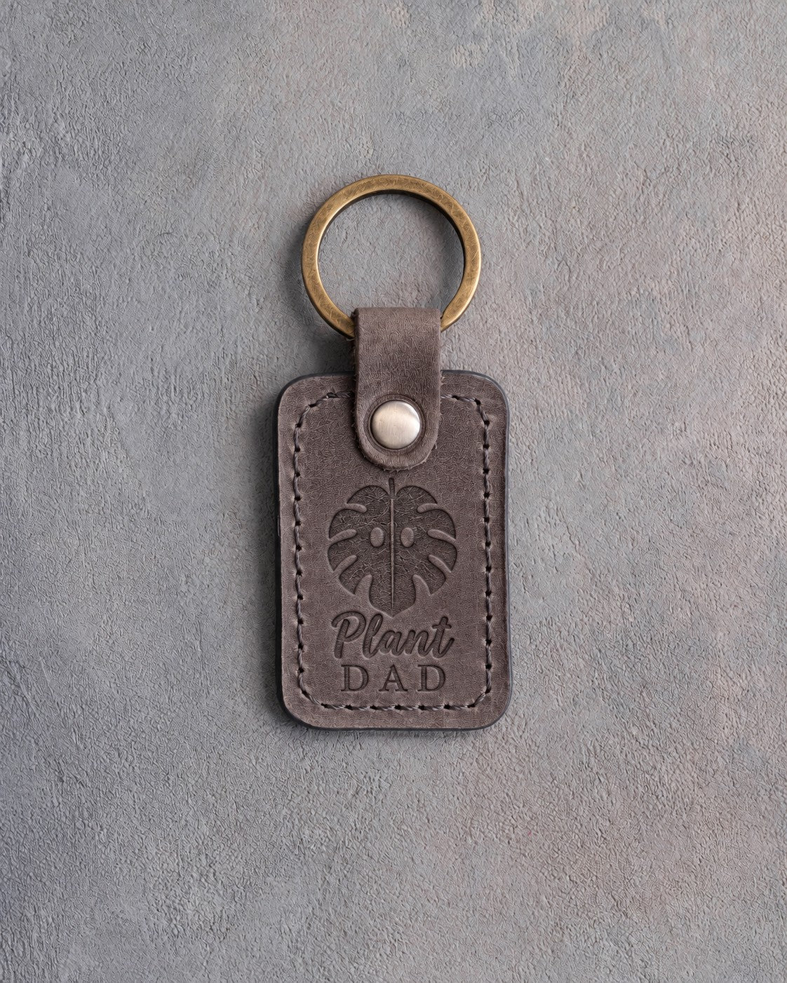 Country gray leather keychain debossed with a monstera leaf above the words Plant DAD, on gray concrete