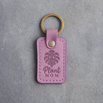 Lilac leather keychain debossed with a monstera leaf above the words Plant MOM, on gray concrete