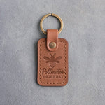 Caramel leather keychain debossed with a honeybee above Pollinator FRIENDLY, on gray concrete