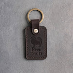Dark espresso leather keychain debossed with a Pomeranian silhouette and the words Pom DAD, on gray concrete