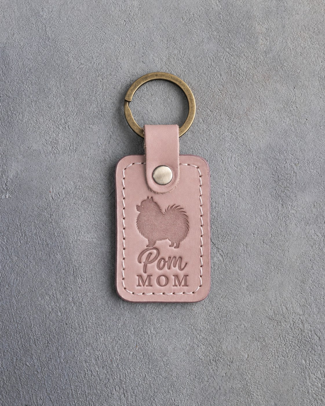 Blush pink leather keychain debossed with a Pomeranian silhouette and the words Pom MOM, on gray concrete