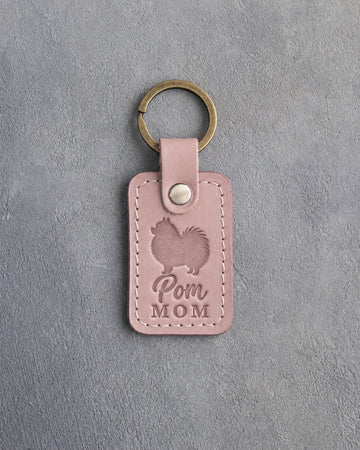 Blush pink leather keychain debossed with a Pomeranian silhouette and the words Pom MOM, on gray concrete