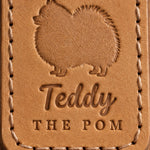 Close-up of Teddy in brush script and THE POM pressed into Indian Summer tan full-grain leather beneath a Pomeranian silhouette