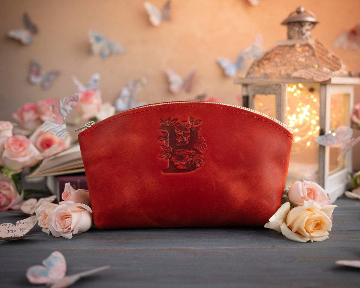 Poppy red initial makeup pouch with a floral monogram, staged among pale roses on a slate table