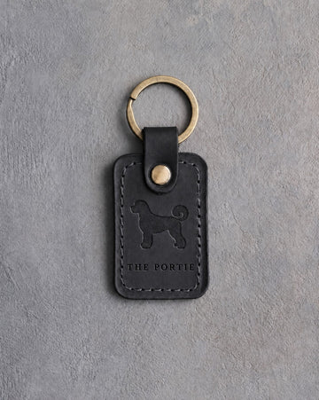 Black leather keychain debossed with a curly Portuguese water dog above THE PORTIE, on gray concrete