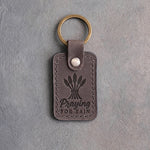 Country gray leather keychain debossed with a tied wheat sheaf above the words Praying FOR RAIN, on gray concrete