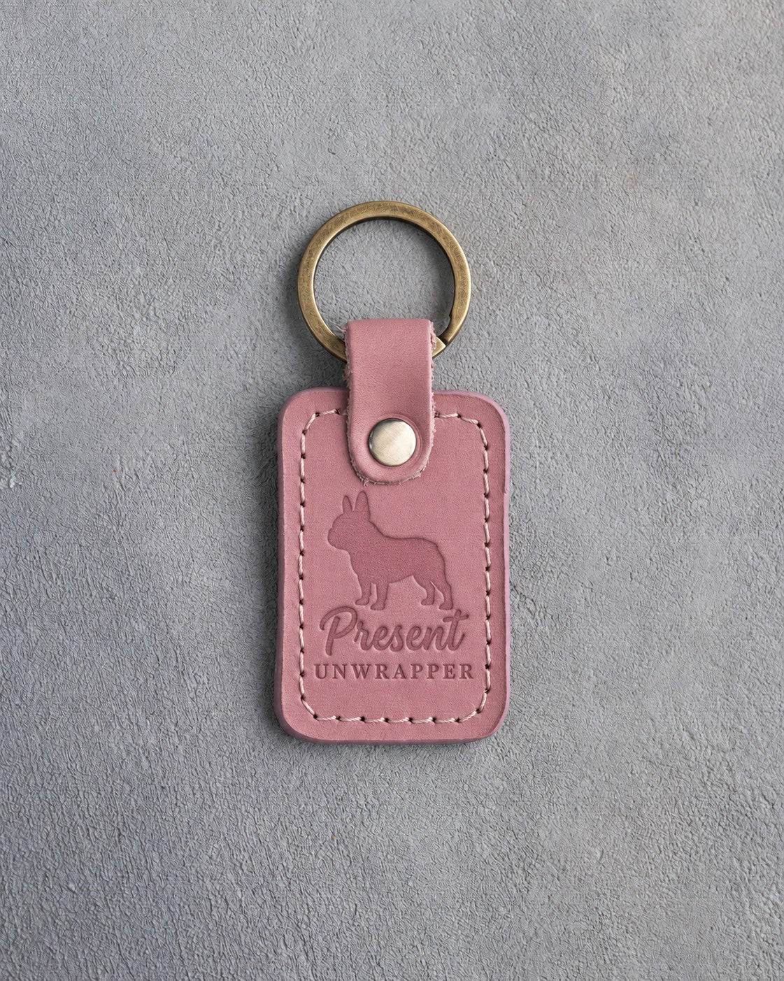 Parisian Blossom pink leather keychain debossed with a French bulldog and the words Present UNWRAPPER, on gray concrete