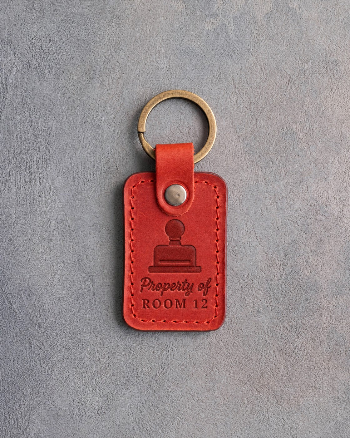 Poppy red leather keychain debossed with a library date stamp above Property of and ROOM 12, on gray concrete