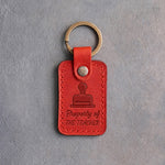 Poppy red leather keychain debossed with a library date stamp above Property of and THE TEACHER, on gray concrete