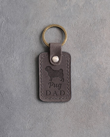 Country gray leather keychain with a debossed pug silhouette and Pug DAD lettering, on gray concrete