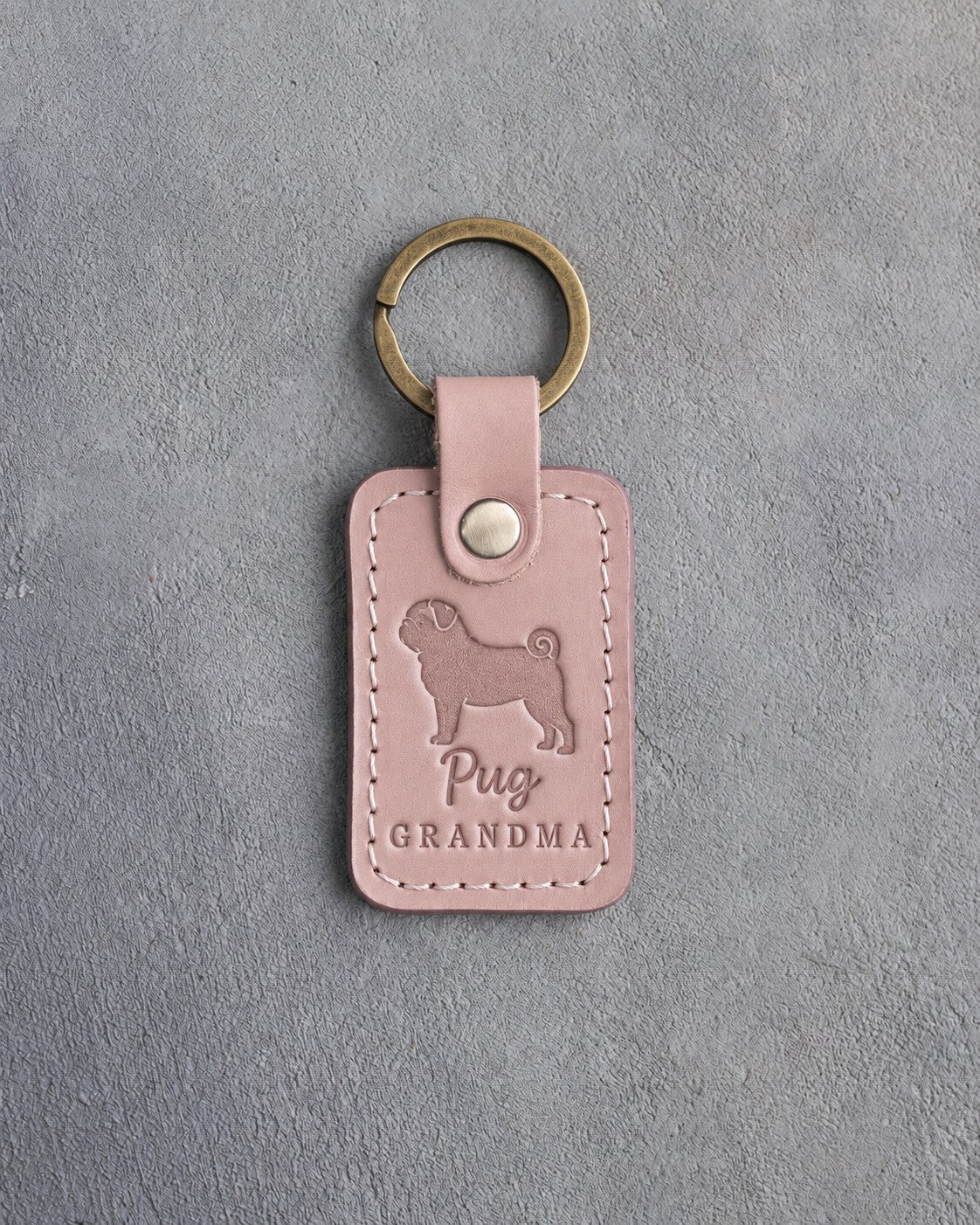 Blush pink leather keychain debossed with a pug silhouette and the words Pug GRANDMA, on gray concrete