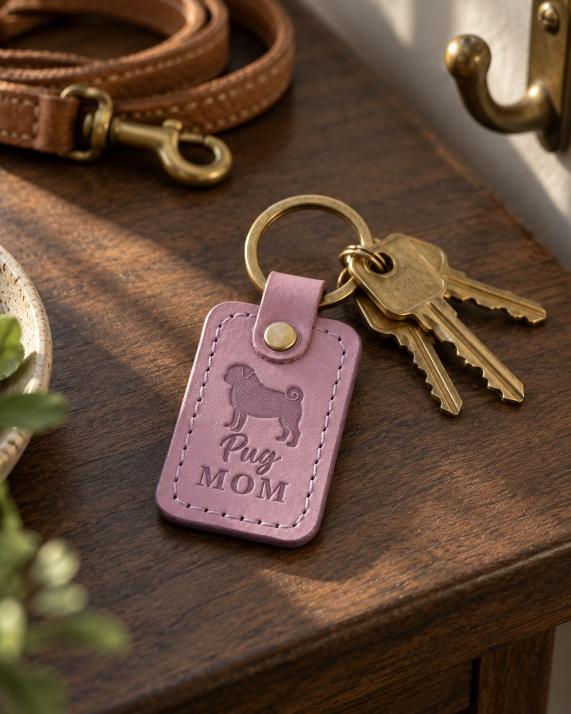 Lilac Pug Mom keychain with brass keys on a dark walnut table, a leather leash and wall hook behind