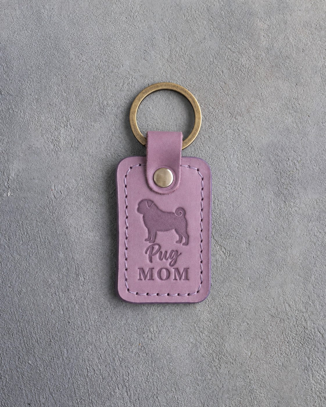 Lilac leather keychain debossed with a pug silhouette and the words Pug MOM, lying on gray concrete