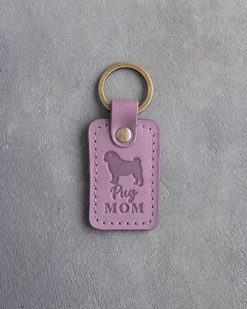 Lilac leather keychain debossed with a pug silhouette and the words Pug MOM, lying on gray concrete
