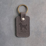 Country gray leather keychain debossed with a cockapoo above Milo and BORN 8·1·26, on gray concrete